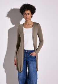 Cardigan lungo olive con frontale drappeggiato, abbinato a una maglietta bianca e jeans a gamba larga blu. Tessuto morbido, maniche lunghe e tasche laterali.