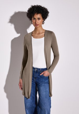 Long gilet olive avec un devant drapé, associé à un haut blanc et un jean large bleu. Tissu lisse, manches longues, et poches latérales.