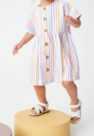 Robe rayée multicolore à manches courtes, boutonnée sur le devant, avec une taille froncée. Associée à des sandales blanches avec des détails festonnés.