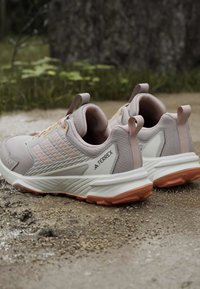 Ζευγάρι ανοιχτό ροζ παπούτσια τρεξίματος Adidas Terrex για μονοπάτια με καφέ σόλες τοποθετημένα σε χαλικόδρομο εξωτερικού χώρου.