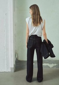 Vrouw met lang haar, gekleed in een witte mouwloze top en zwarte wijde jeans, met een bijpassende zwarte spijkerjack in haar hand, staand binnen.