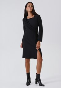 Zwarte jurk met lange mouwen, gedrapeerde halslijn en een zijopening. Gecombineerd met strakke zwarte enkellaarsjes. Glad materiaal, getailleerd silhouet.