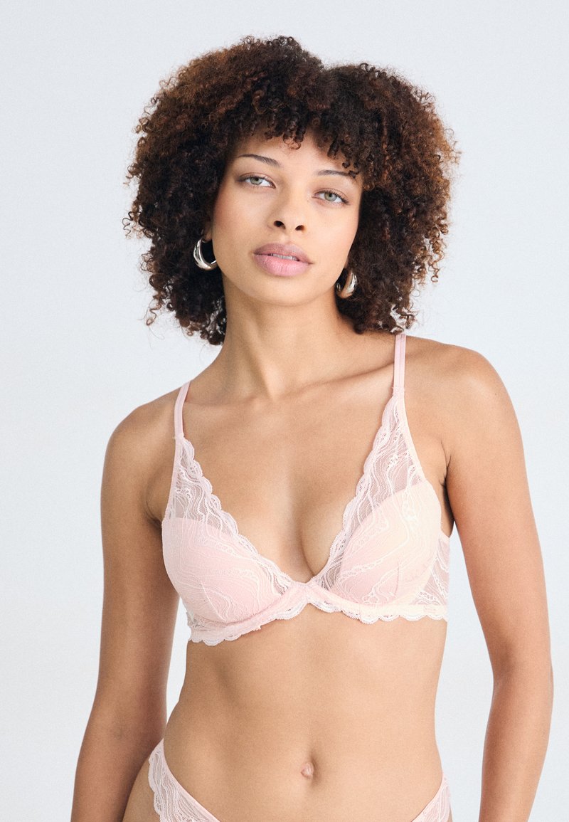Soutien-gorge en dentelle rose avec des bords festonnés, des bonnets rembourrés et des bretelles ajustables, présentant un motif floral et des détails délicats le long de la bande.