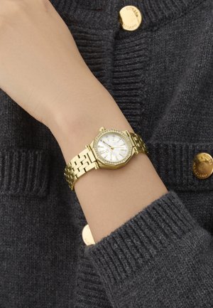 Auksinis aštuonkampis laikrodis su metaliniu apyrankiu ir baltu ciferblatu, su kristaliniais akcentais aplink bezelį, uždėtas ant rankos su pilku megztiniu.