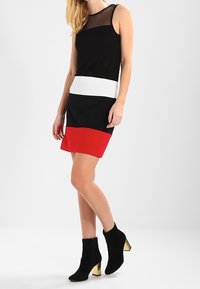 Vestido negro sin mangas con parte superior de malla, franja horizontal blanca y panel inferior rojo. Par de botas negras hasta el tobillo con acentos dorados en los tacones.