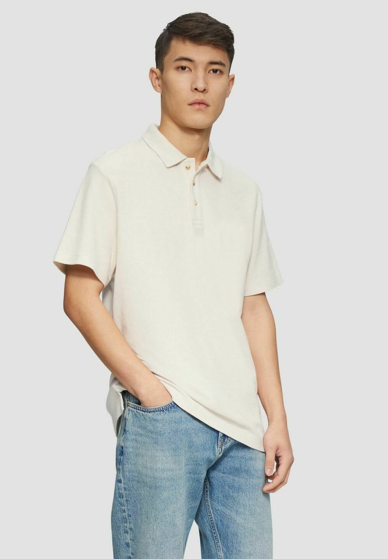 Helles beiges Poloshirt mit kurzen Ärmeln, Knopfleiste mit drei Knöpfen und lockerer Passform, getragen mit blauen Jeans. Glatte Textur, minimalistisches Design.