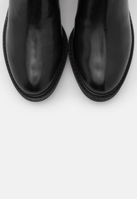Botas de tobillo de cuero negro con un acabado brillante, punta redondeada y detalle cosido alrededor de la suela. Textura suave y sin adornos.