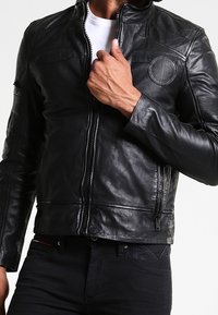 Schwarze Lederjacke mit taillierter Passform, Reißverschluss, runden und strukturierten Akzenten an den Schultern sowie dezenten Steppdetails.