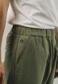 Olivové kalhoty se elastickým pasem, bočními kapsami a jemným brandingem. Vyrobeno z měkké, strukturované tkaniny. Pohodlný střih.