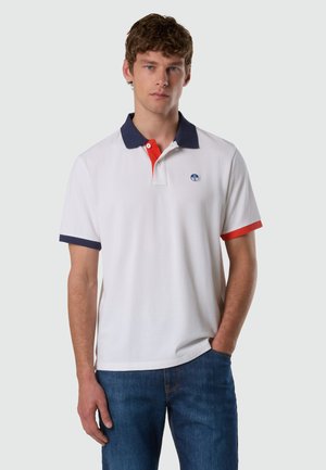 Giovane uomo indossa una polo bianca con collo e bordi delle maniche blu navy e rossi, abbinata a jeans blu, in piedi contro uno sfondo neutro.