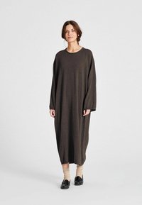 Ženska stoji oblečena v ohlapen, dolg temno rjav plašč, bež nogavice in črne natikače na enotnem svetlem ozadju.