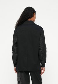 Kvinne med langt krøllete hår som har på seg en svart blazer med rynkede ermer og svarte cargo-bukser, vendt bort fra kamera mot en hvit bakgrunn.