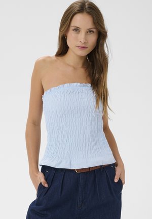 Jeune femme aux longs cheveux bruns portant un haut smocké bleu clair sans bretelles et un pantalon taille haute bleu foncé, les mains dans les poches.