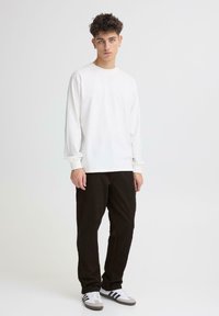 Camiseta blanca de manga larga y cuello redondo combinada con pantalones negros. El modelo lleva zapatillas blancas y grises con rayas negras. Textura lisa y uniforme.