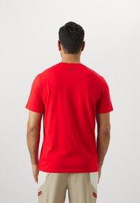Rød kortærmet t-shirt lavet af blødt stof, med rund hals og et lille sort logo på bagsiden, båret med beige shorts.