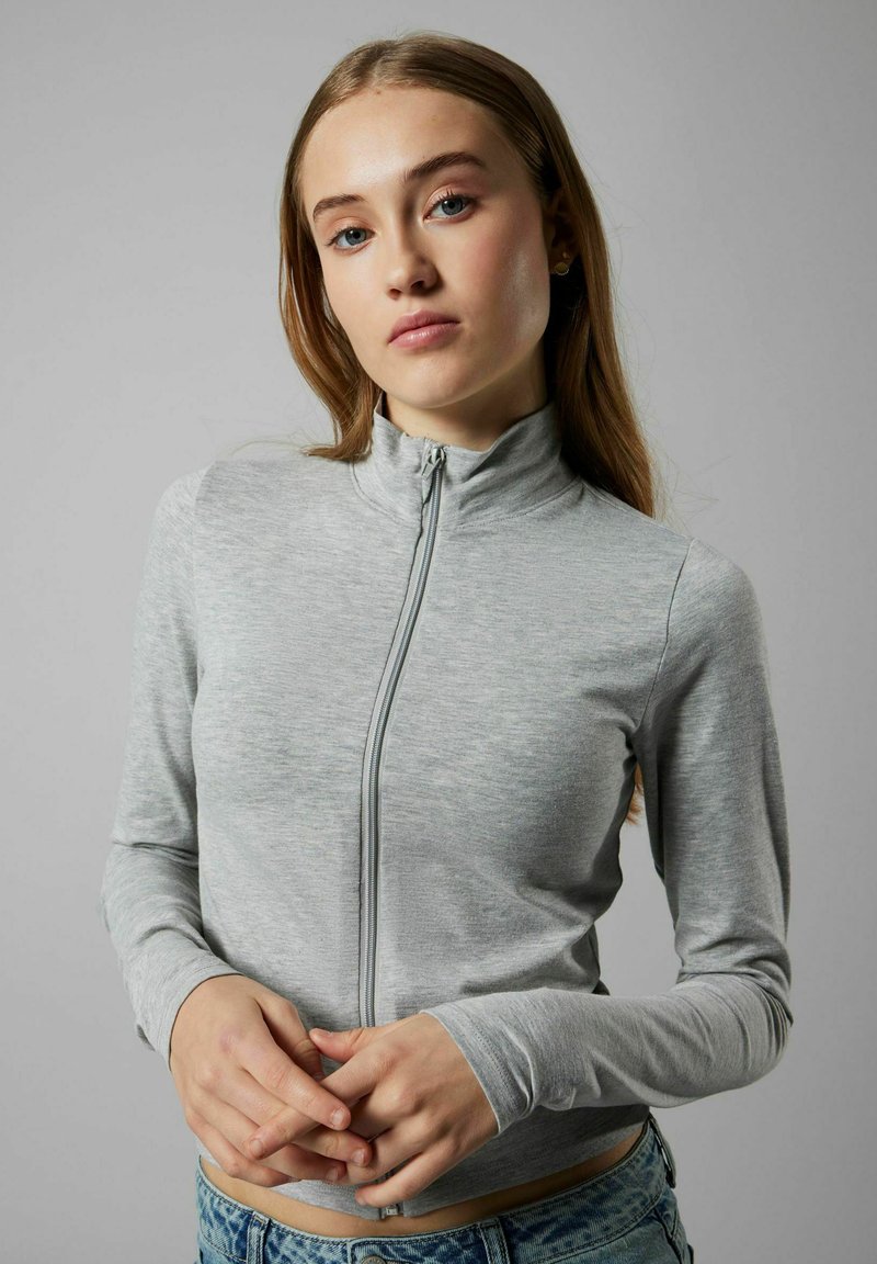 Haut zippé à manches longues gris clair, fabriqué à partir d'un matériau lisse et extensible, avec un col haut et une coupe ajustée. Tissu texturé visible.