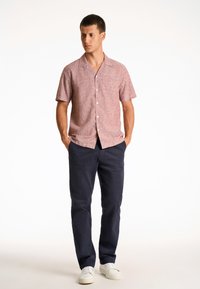 Camisa de manga corta y botones en una tela roja con un patrón sutil, combinada con pantalones azul marino y zapatillas blancas. El modelo está de pie recto.