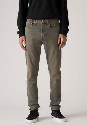 Mann trägt ausgewaschene olivgrüne Slim-Fit-Jeans, schwarzes Langarmshirt und schwarze Sneakers mit weißen Sohlen, steht vor einfachem Hintergrund.