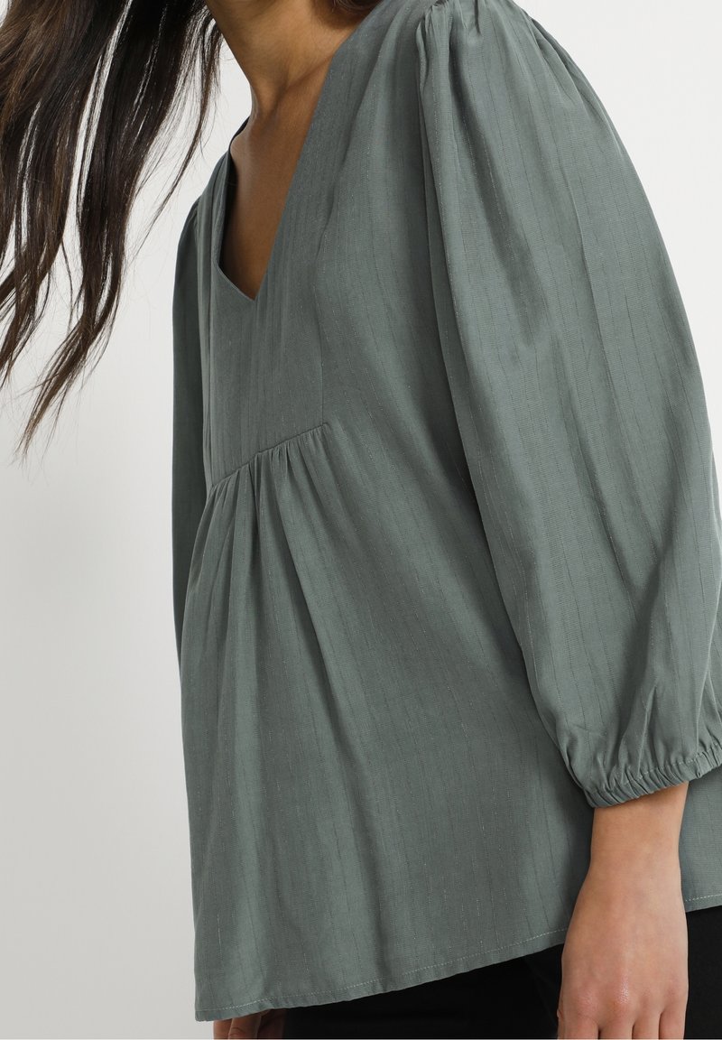 Femme portant un chemisier vert ample à manches longues, avec un profond décolleté en V et un tissu froncé sous la poitrine, debout devant un fond uni.