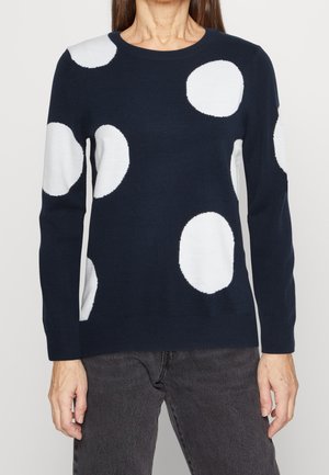 Femme portant un pull bleu marine avec de grands pois blancs et un jean gris foncé, debout devant un fond clair uni.