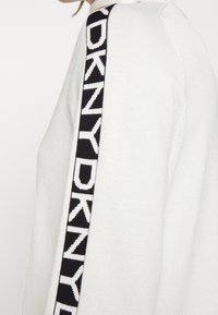 Vit stickad tröja med svart sidstripe och "DKNY"-text i vitt, fin textur och figurnära design.