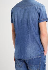 Camicia in denim a maniche corte di blu medio, con un design a yoke appuntito, collare piatto e taglio dritto con sfumature leggere e dettagli di cucitura.