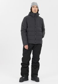 Schwarze Daunenjacke mit durchgehendem Reißverschluss, Kapuze und erhöhtem Kragen. Kombiniert mit lockeren schwarzen Hosen und schwarzen Winterstiefeln. Graue Mütze mit Logo.