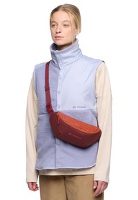 Lichtblauwe fleece vest met knoopsluiting, gecombineerd met een crèmekleurig longsleeve shirt en een rood-oranje schoudertas met logo-details.