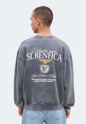 Άνδρας με πλάτη φοράει γκρι φούτερ SL Benfica με λογότυπο αετού και λευκό κείμενο, συνδυασμένο με ανοιχτόχρωμα μπλε τζιν.