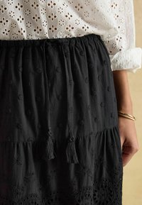 Czarna bawełniana spódnica z haftowanymi kwiatowymi wzorami, warstwowym fasonem, sznurkami do wiązania z frędzlami oraz koronkową lamówką na dole.