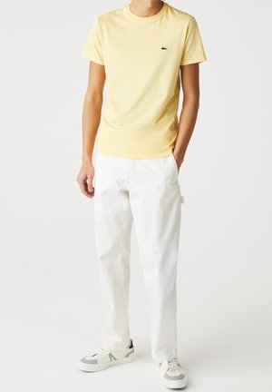 T-shirt en coton jaune avec un col rond et un petit logo Lacoste, associé à un pantalon blanc ample et des baskets grises avec des accents verts.