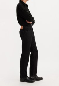 Personne debout, les bras croisés, portant un t-shirt noir à manches longues, un jean noir avec un patch marron et des bottes en cuir noir sur fond blanc.