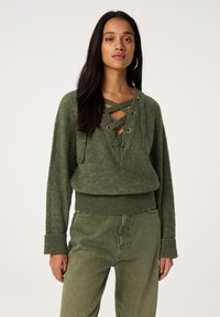 Jonge vrouw met lang donker haar, grote creolen, een olijfgroen gebreide trui met veters en bijpassende broek met hoge taille, tegen een witte achtergrond.