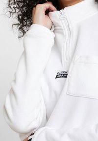 Vit zip-sweatshirt med kvartdragkedja, tillverkad av mjukt material, med en bröstficka framtill och en svart Adidas-logotyp patch på vänster sida.