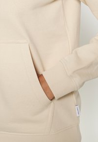 Felpa in cotone beige con polsini a costine, tasca a marsupio e piccolo etichetta sul lato. Tessuto morbido e finitura liscia.