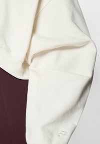 Kremfarget sweatshirt med ribbestrikkede mansjetter og rå kant. Myk tekstur på overarmen med en liten logo nær mansjetten. Burgunder bakgrunn.