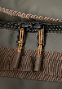 Green fabric zipper section with brown nylon trim and black hardware. Features two golden cord pulls with textured grips for easy opening.