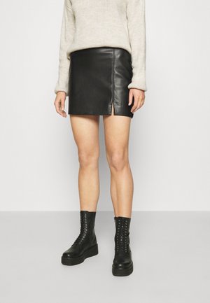 Personne portant un pull crème, une mini-jupe en cuir noire avec une fente latérale, et des bottes à plateforme noires lacées, debout devant un fond uni.