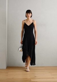 Black sleeveless dress with a draped neckline, asymmetrical hem, and metallic strap accents. Paired with a silver handbag and white sandals.