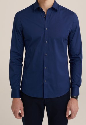 Man wearing fitted long-sleeve navy blue button-up shirt with collar and black wristwatch, standing against plain beige background.