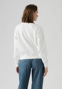 Felpa bianca in cotone con collo tondo, polsini e orlo a coste, indossata con jeans in denim blu a vestibilità ampia, mostrata da dietro.