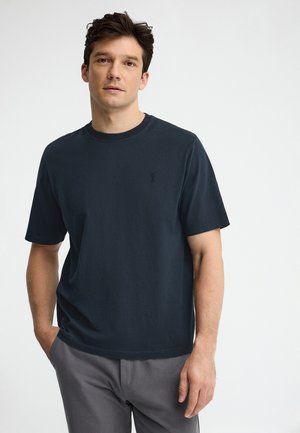 Homme aux cheveux foncés portant un t-shirt à manches courtes bleu marine et un pantalon gris, debout devant un fond clair uni.