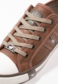 Baskets marron avec lacets beiges, bout en caoutchouc blanc, œillets métalliques et logo cheval mustang sur le lacet et la semelle.