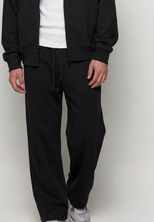 Mann trägt schwarze Sweatpants, weiße Sneaker, schwarze Zip-up-Jacke und weißes T-Shirt vor einem schlichten hellen Hintergrund.