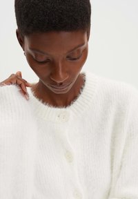 Weißer Cardigan mit weicher, flauschiger Textur, Knopfverschluss und gerippten Kanten. Verfügt über einen runden Ausschnitt und sichtbare Nähte.