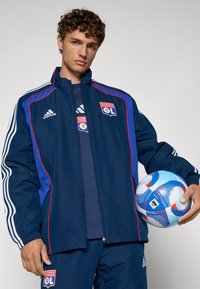 Marineblaue Sportjacke mit blauen und roten Akzenten, Reißverschlüssen und Logos. Das Modell hält einen mehrfarbigen Fußball und trägt passende Sportbekleidung.