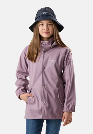 Dziewczyna ubrana w jasnofioletową wodoodporną kurtkę i granatowy kapelusz typu bucket, stojąca z jedną ręką w kieszeni kurtki na jednolitym tle.