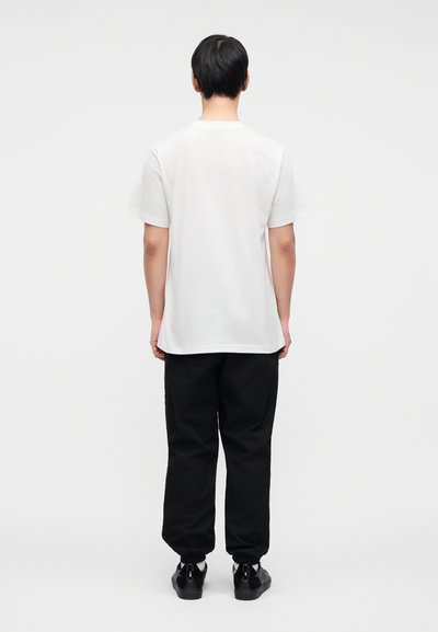 Λευκό κοντομάνικο T-shirt που φαίνεται από πίσω, συνδυασμένο με μαύρο παντελόνι με στενή γραμμή κ αι μαύρα παπούτσια. Απλός σχεδιασμός, χωρίς ορατά μοτίβα ή λογότυπα.