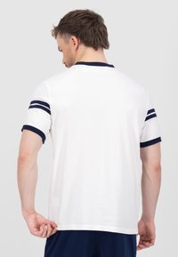 Balts īsām piedurknēm t-krekls ar tumši zilu ribotu apkakli un piedurkņu joslām, izgatavots no gludas, vieglas auduma. Aizmugures skats rāda vienkāršu dizainu.