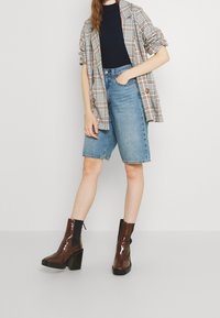 Blazer oversize a quadri, top aderente nero, shorts in denim a vita alta e stivali alla caviglia marroni lucidi con tacco spesso.